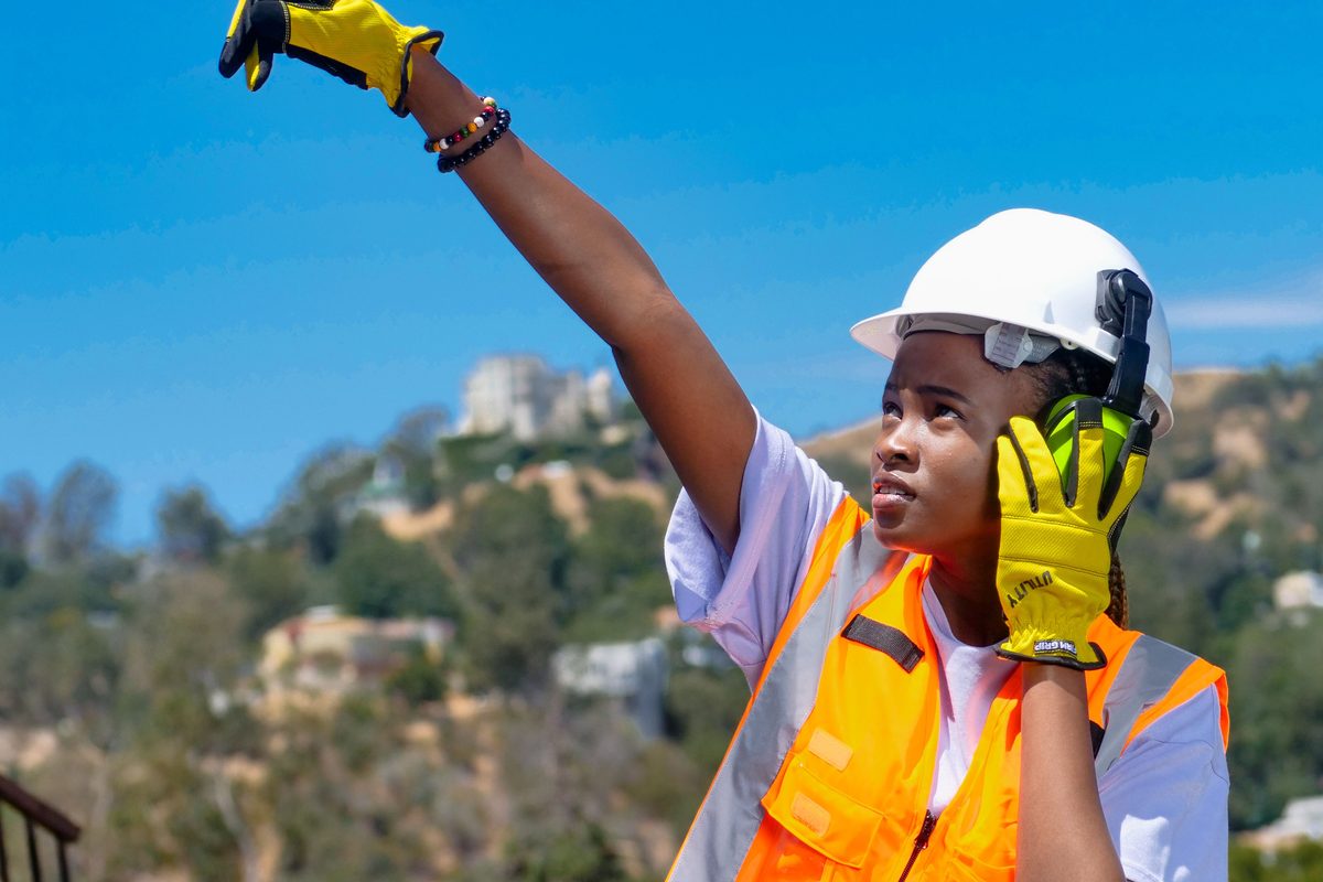 A site worker in a hard hat and orange hi-vis vest signalling with a raised arm while holding a radio