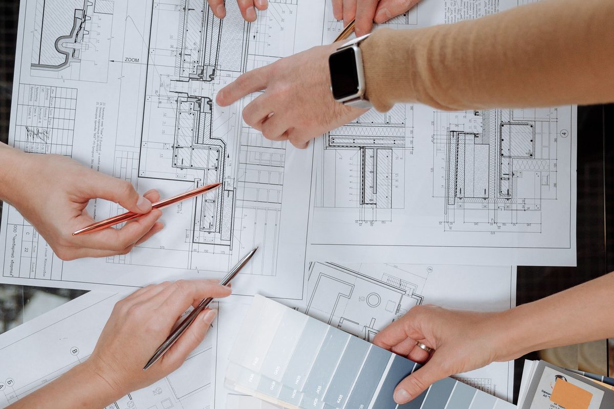 Four pairs of hands over overlapping floor plans, one pointing at a detail while another holds a card of finish samples