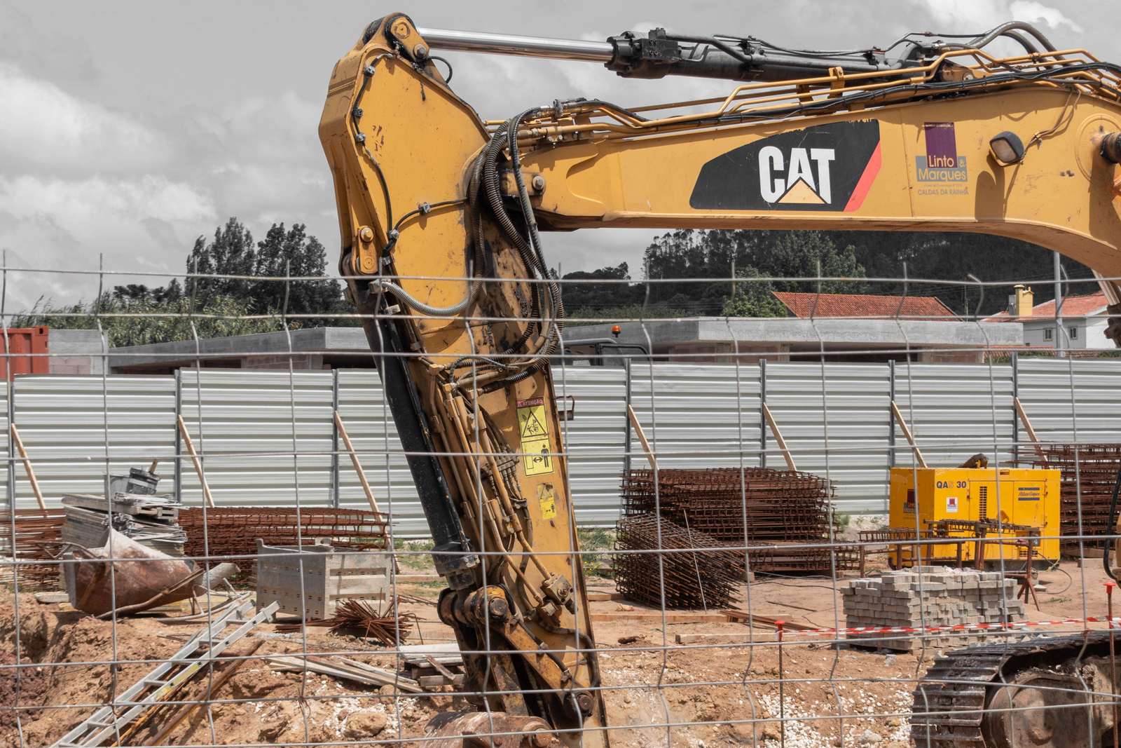 A tracked excavator standing over a fenced foundation site, with stacked reinforcement cages waiting beside the open trench