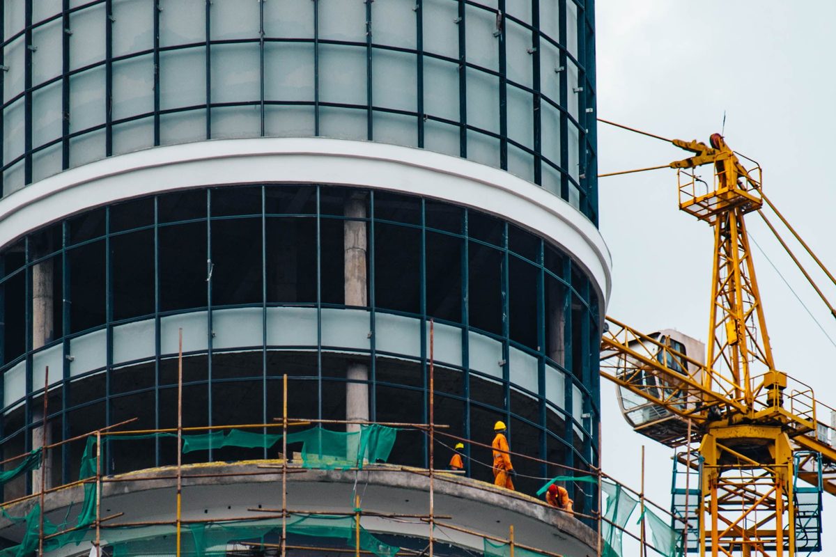 A curved glass-clad tower still in scaffold, with a tower crane alongside and workers on the platform at its base