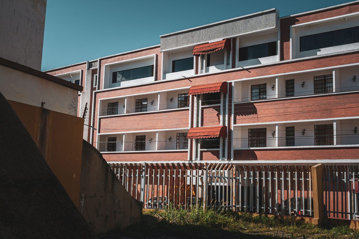 A four-storey block with brick infill panels and shaded balcony awnings, seen from the road behind a steel perimeter fence