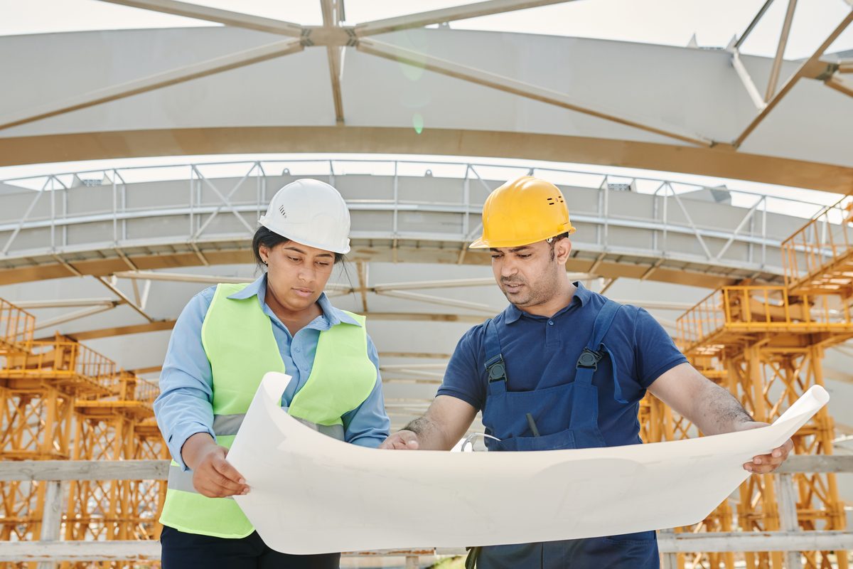 Two site staff in hard hats holding an unrolled drawing open between them, with yellow falsework towers rising behind