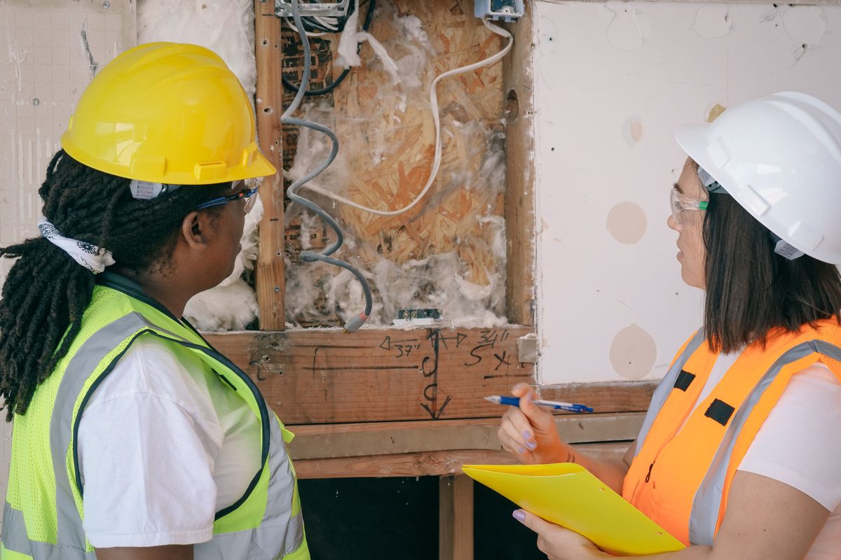 Two surveyors in hard hats examining an opened-up wall cavity, the exposed timber marked with pencil annotations